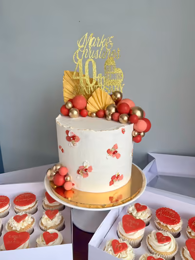 A white buttercream anniversary cake with red and gold abstract floral details, topped with a 'Mark & Christine 40th Anniversary' gold glitter topper, gold fan decorations, and clusters of red and gold spheres. The cake is surrounded by boxes of matching cupcakes with white frosting and red heart and 'Happy Anniversary' toppers.