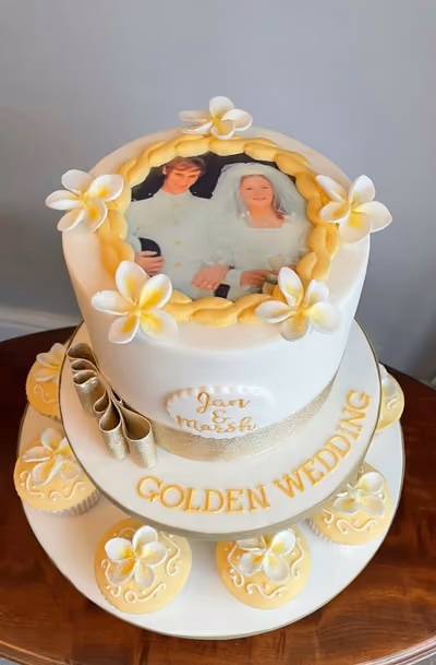 A two-tier white cake celebrating a golden wedding anniversary, featuring a framed wedding photo of the couple on the top tier, gold ribbon, white and yellow sugar flowers, and text that reads 'Jan & Marsh' and 'GOLDEN WEDDING'. The cake is surrounded by matching yellow cupcakes with white and yellow flowers.