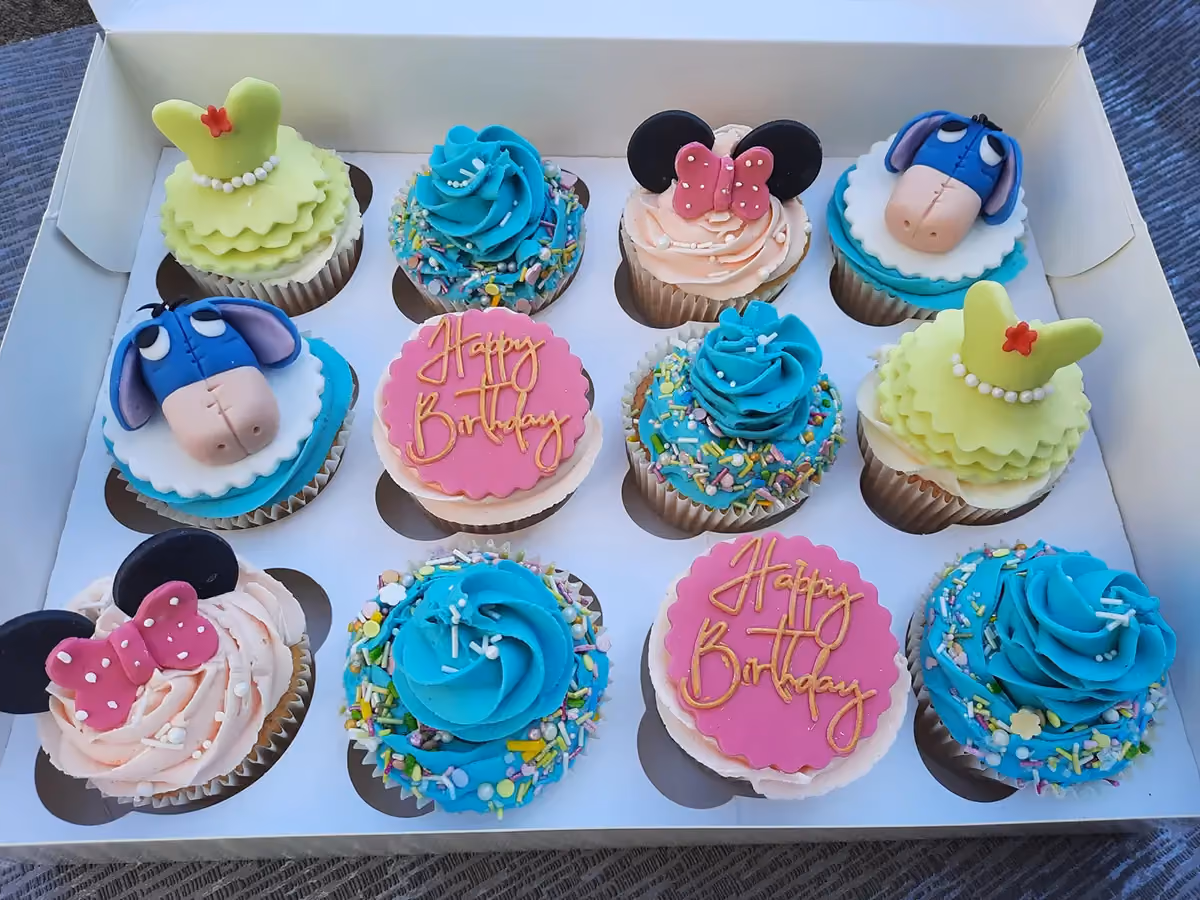 A dozen assorted themed cupcakes in a white box, featuring designs of Minnie Mouse, Eeyore, green princess dresses, blue frosting with colorful sprinkles, and pink 'Happy Birthday' toppers.