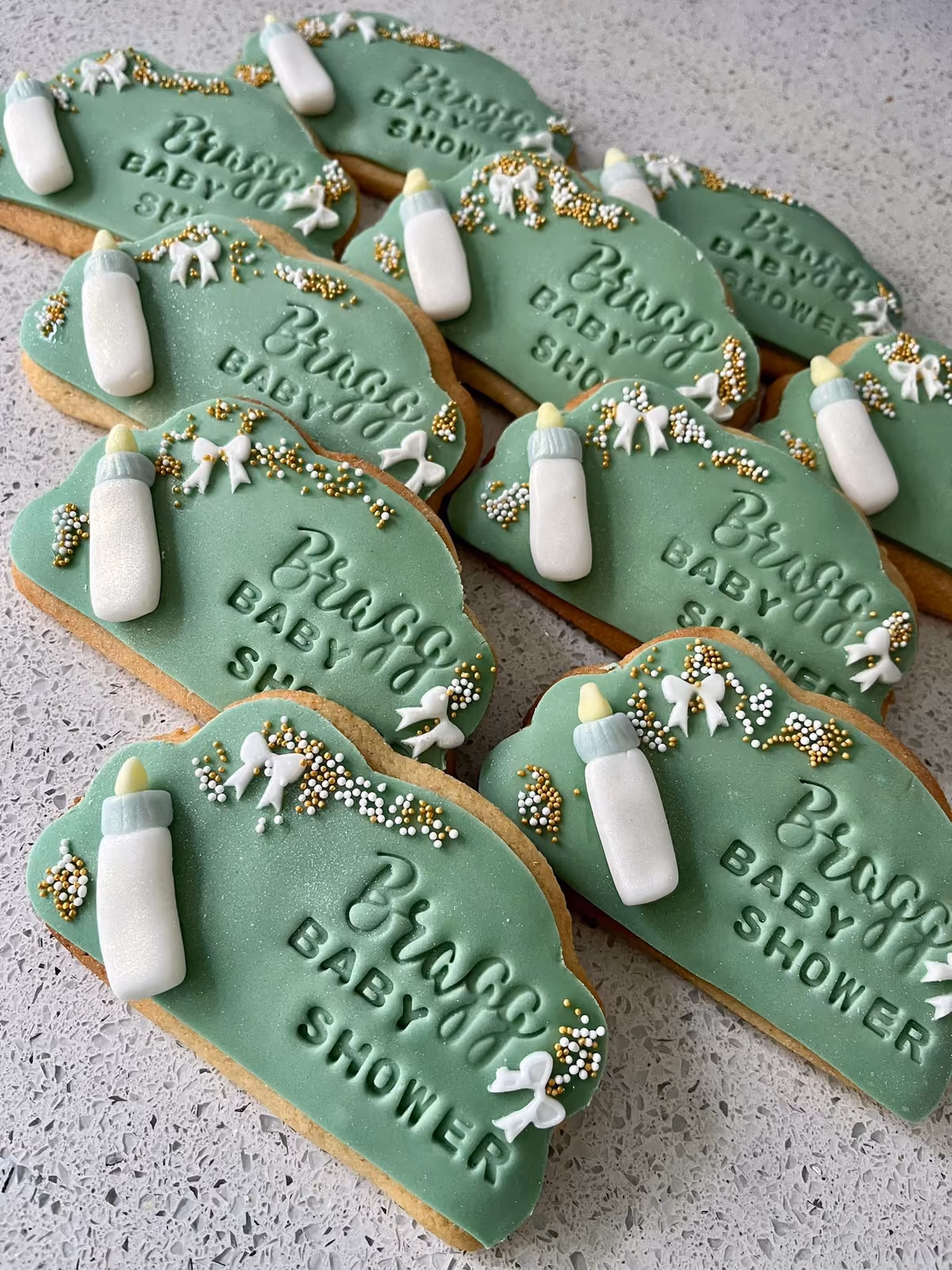 Close-up of multiple green plaque-shaped baby shower cookies, each featuring a white baby bottle, white fondant bows, gold and white sprinkles, and the embossed text 'Braggs BABY SHOWER'.