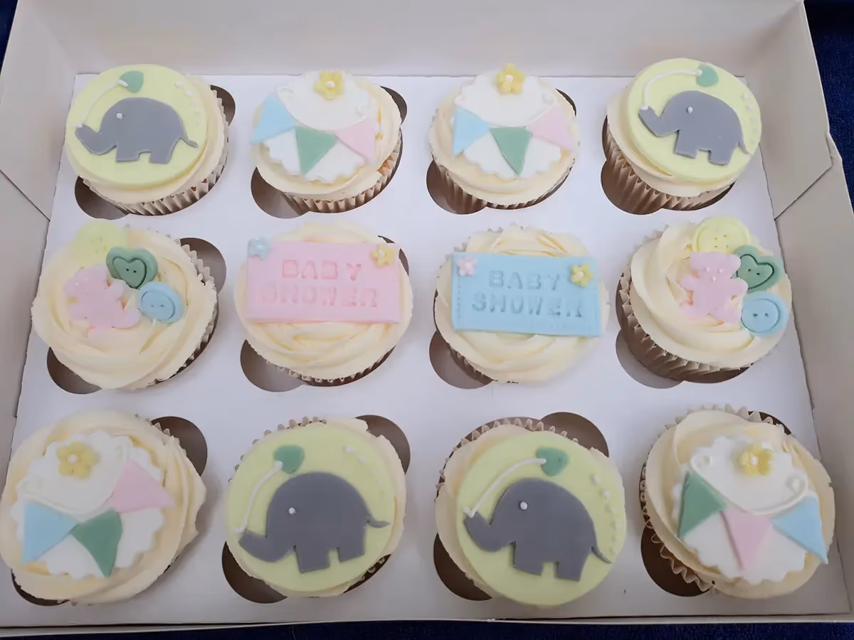 A dozen baby shower cupcakes with white frosting, arranged in a white box, featuring assorted pastel fondant decorations. Designs include grey elephants with green balloons, banners with blue, pink, and green flags, 'Baby Shower' text on pink and blue plaques, and a mix of baby-themed shapes like pink teddy bears, green hearts, and yellow and blue buttons.