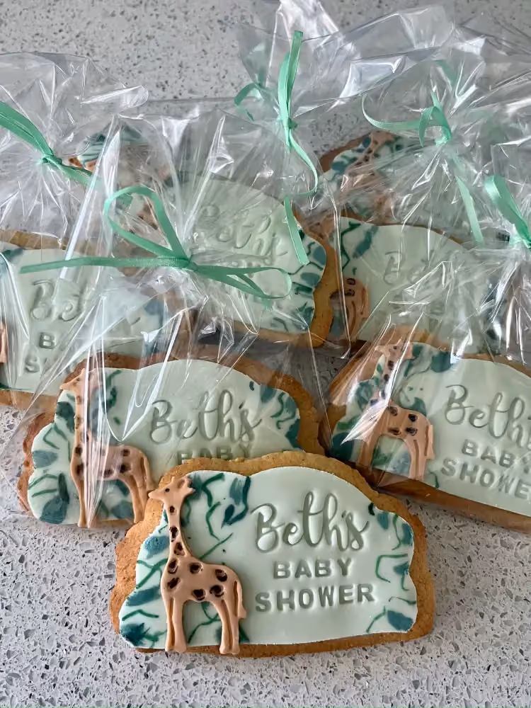 Several individually packaged baby shower cookies, each decorated with light green and white icing, a giraffe figure, and the text 'Beth's BABY SHOWER', tied with a mint green ribbon.