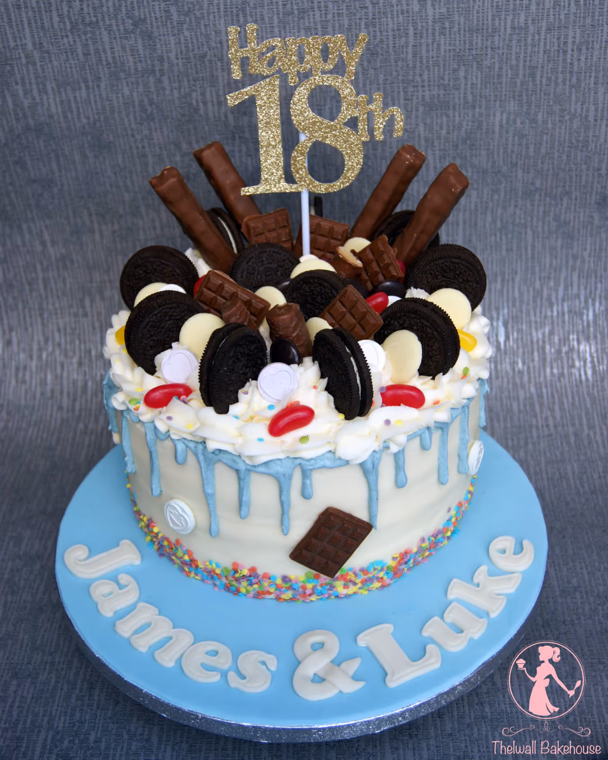 An 18th birthday drip cake for James and Luke, decorated with a variety of chocolates, Oreo biscuits, white chocolate discs, and jelly beans, featuring a gold 'Happy 18th' topper, white frosting with blue drips, and colorful sprinkles on a light blue cake board.