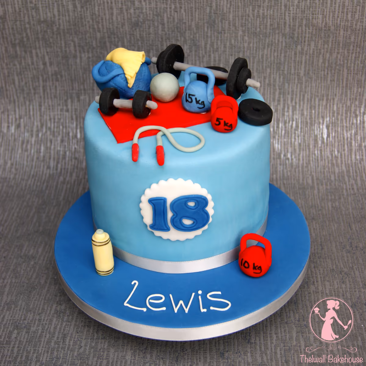 A light blue fondant birthday cake for an 18th birthday, decorated with various gym and fitness-themed sugar craft items including dumbbells, kettlebells, a gym bag, and a skipping rope, with the name 'Lewis' written on the blue cake board.