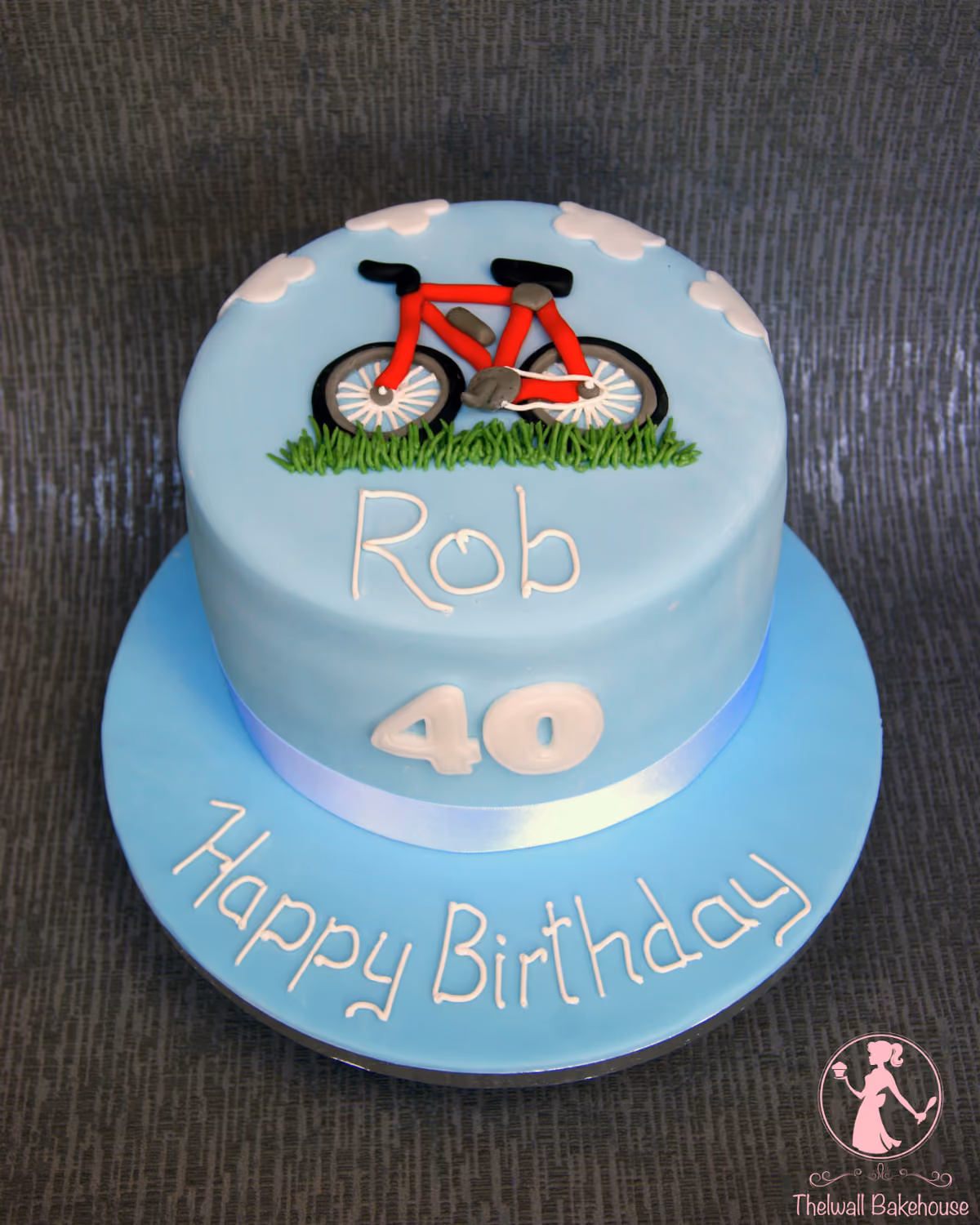 A light blue round birthday cake for Rob's 40th, decorated with a red bicycle, green grass, and white clouds on top, with 'Happy Birthday' written on the cake board.