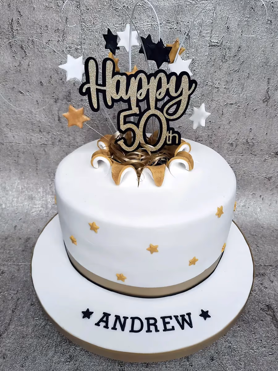 A white fondant 50th birthday cake with a gold band, decorated with gold stars and a "Happy 50th" gold glitter topper. The cake board is personalized with the name "ANDREW" in black letters and black stars.