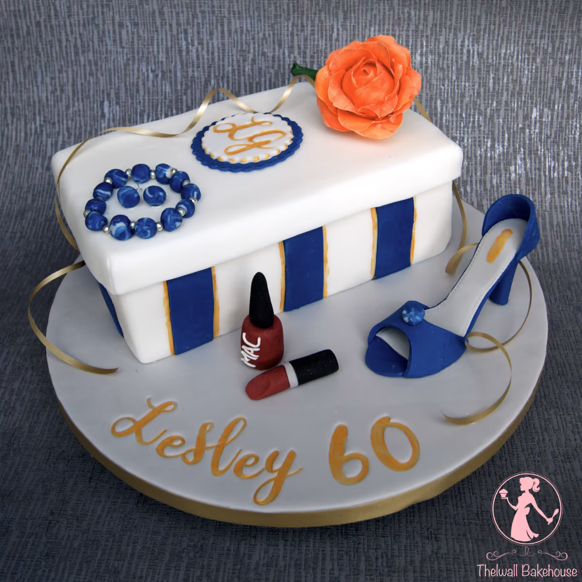 A white gift box shaped birthday cake for Lesley's 60th, featuring blue and gold stripes, an orange sugar rose, blue beaded jewelry, a blue high-heeled shoe, red nail polish, and lipstick, with 'Lesley 60' written in gold on the white cake board.