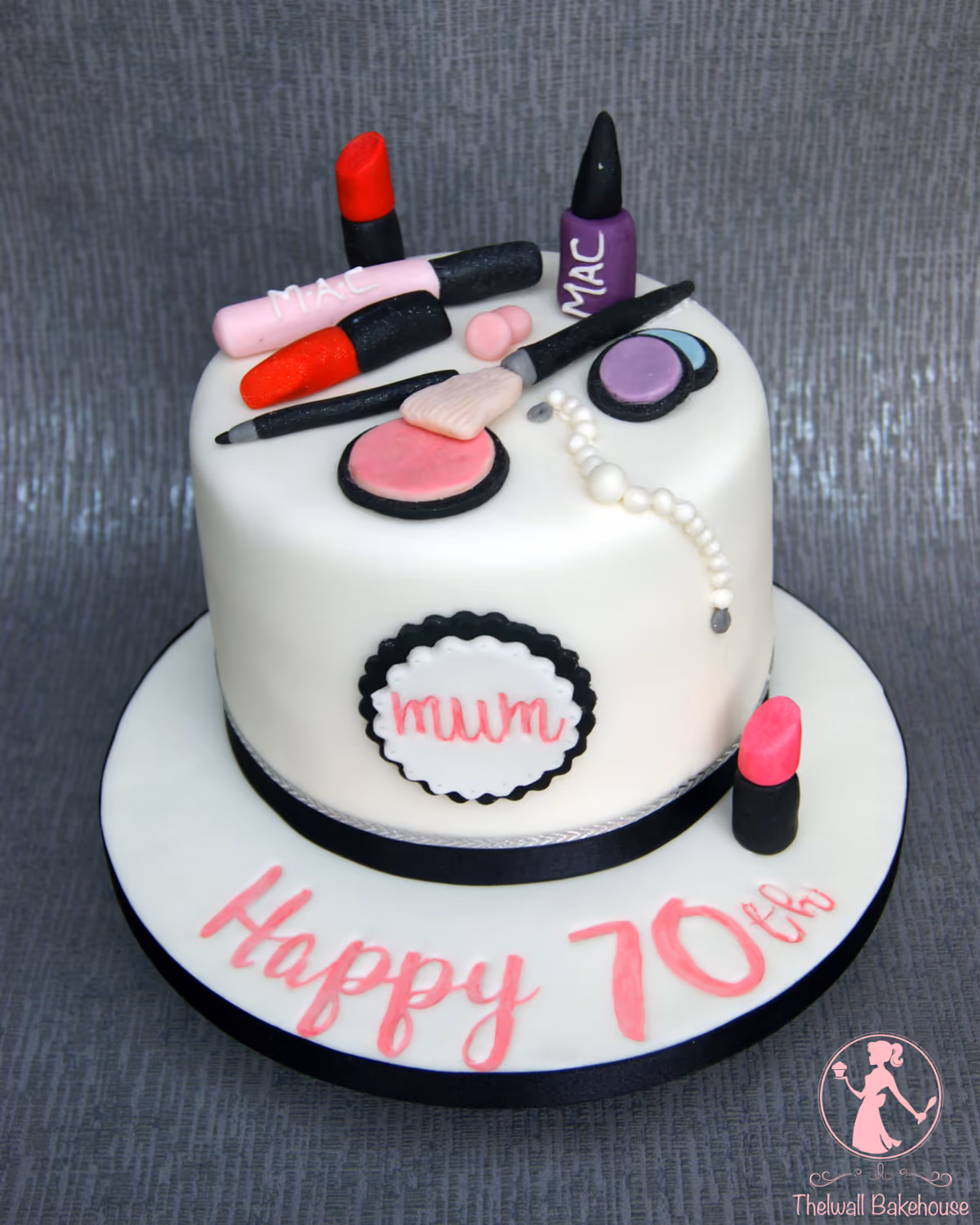 A round white fondant cake decorated with various edible makeup items, including lipsticks, nail polish, eyeshadow, and brushes, along with a string of pearls. The cake features a 'mum' inscription on the side and 'Happy 70th' written in pink on the cake board.
