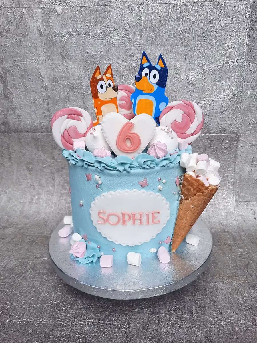 A light blue single-tier birthday cake featuring Bluey and Bingo character toppers, a white heart with a pink number 6, and the name 'SOPHIE' on the side. It is decorated with pink and white lollipops, meringues, marshmallows, and an ice cream cone filled with marshmallows.