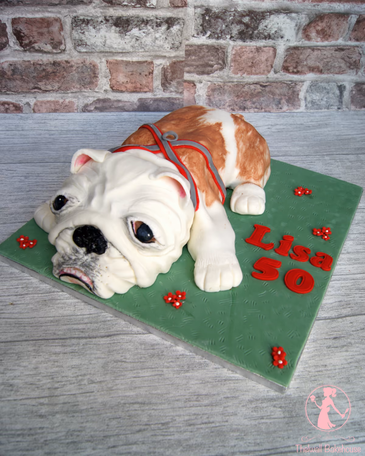 A white and brown bulldog-shaped birthday cake, wearing a red harness, on a green base decorated with red letters spelling 'Lisa 50' and small red flowers.