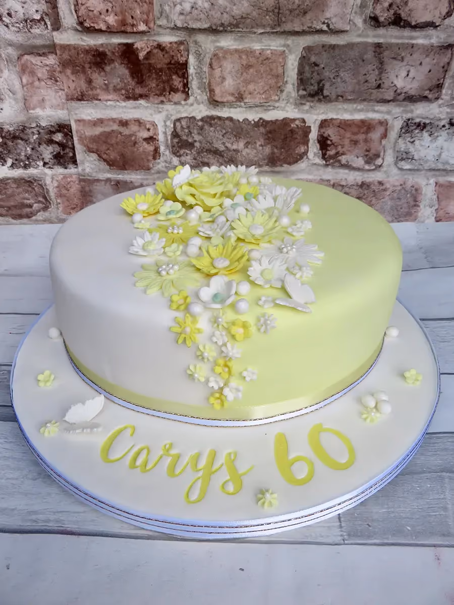 A round birthday cake for Carys' 60th, half covered in white fondant and half in lime green fondant, decorated with a cascade of yellow and white sugar flowers, pearls, and small butterflies. The cake board has 'Carys 60' written in yellow letters and numbers.