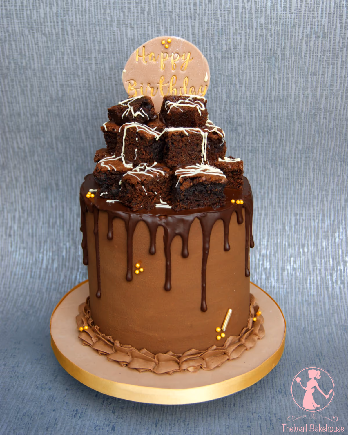 A chocolate birthday cake with smooth chocolate buttercream, a dark chocolate ganache drip, and a large pile of brownie squares drizzled with white icing on top. It features a round 'Happy Birthday' plaque and is decorated with gold sprinkles.