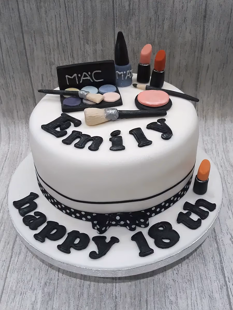 A white round birthday cake decorated with black fondant letters spelling 'Emily' on top, surrounded by makeup-themed decorations including a MAC eyeshadow palette, lipsticks, nail polish, and brushes. The base of the cake features black fondant letters spelling 'happy 18th' and a black and white polka dot ribbon with a bow.