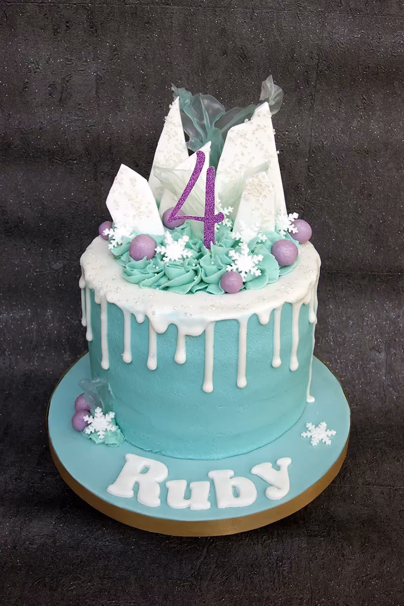 A Frozen-themed birthday cake for Ruby's 4th birthday, featuring a teal base, white drip icing, and topped with white chocolate shards, purple candies, and snowflakes, with a glittery purple number 4.