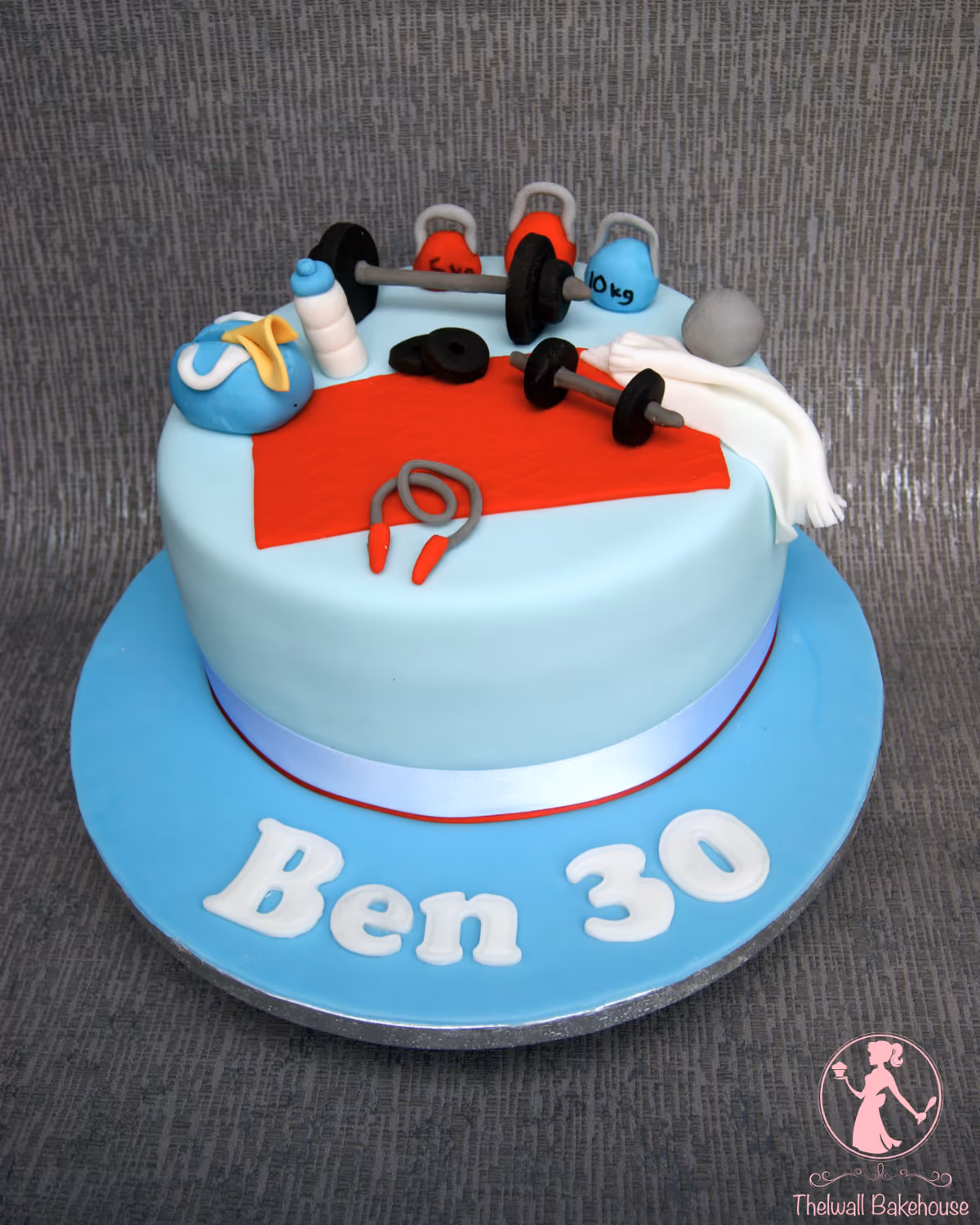 A light blue round birthday cake decorated with various gym and fitness-themed items including barbells, kettlebells, a water bottle, a gym bag, a red yoga mat, a white towel, and a jump rope. The cake board has "Ben 30" written in white letters.