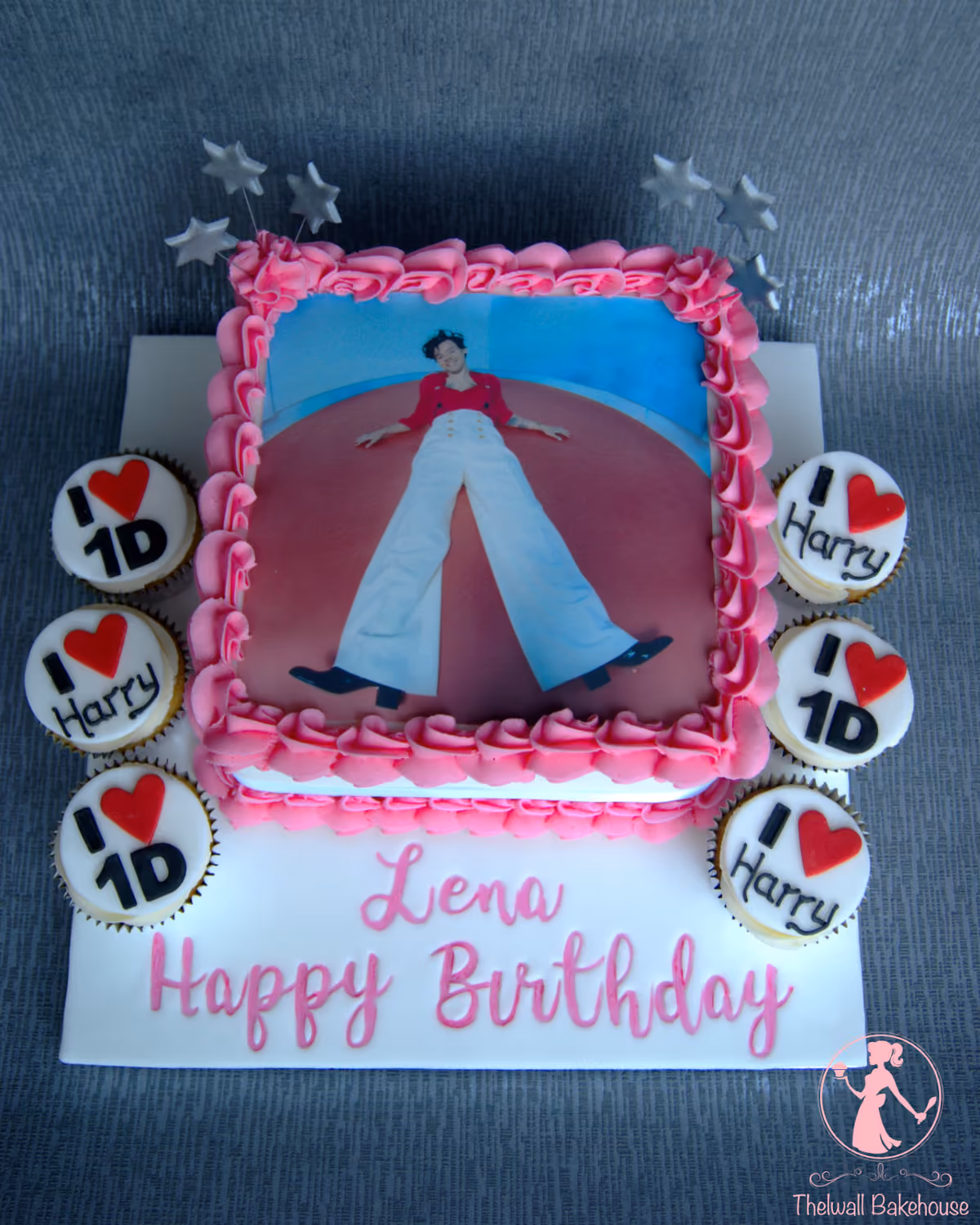 A rectangular white birthday cake featuring a large edible image of Harry Styles, framed by pink piped icing, surrounded by cupcakes decorated with 'I love 1D' and 'I love Harry' messages. The cake board reads 'Lena Happy Birthday' in pink script, and silver star decorations adorn the top of the main cake.