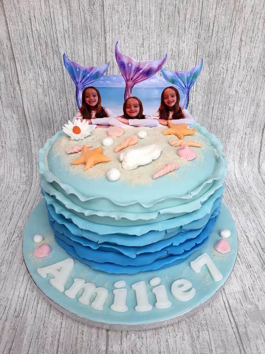 A two-tier mermaid-themed birthday cake featuring blue ombre ruffles on the bottom tier, edible sand, seashells, and orange starfish on the top tier, and three photo toppers of girls with mermaid tails. The light blue cake board reads 'Amilie 7' in white letters.