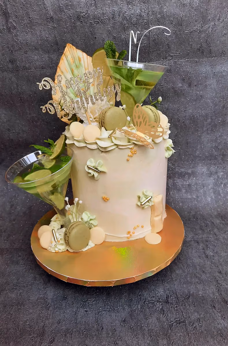 Single-tier birthday cake in light green and cream, decorated with two martini glasses containing lime and mint, green and cream macarons, a gold butterfly, a 'Happy Birthday' topper, and gold sprinkles.