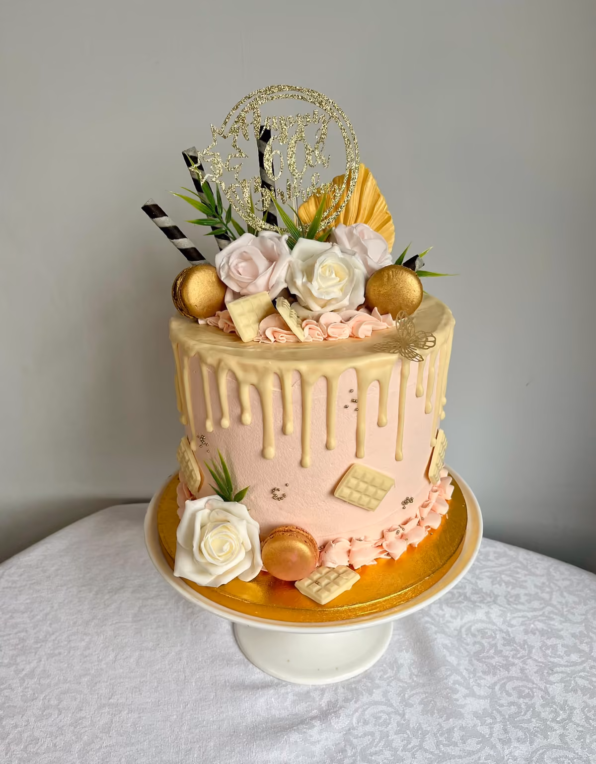 A tall pink birthday cake with a white chocolate drip, decorated with white and pink roses, gold macarons, white chocolate pieces, striped straws and dried palm leaves, with a gold glitter topper.