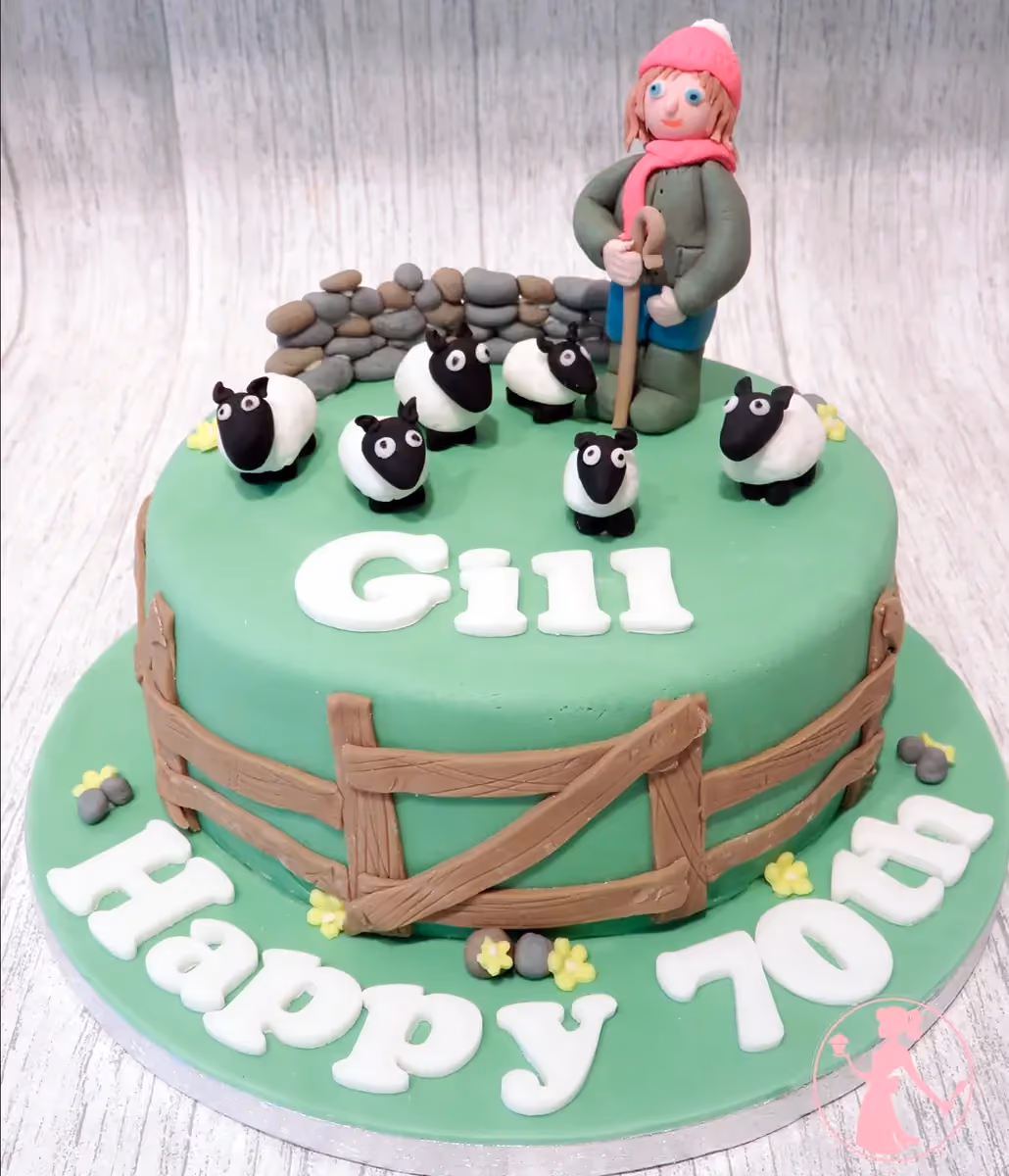 A round green birthday cake decorated with a fondant figure of a shepherdess in a pink hat and scarf, five white and black sheep, a stone wall, and a wooden fence. The name 'Gill' is written on the top and 'Happy 70th' on the base, surrounded by small yellow flowers and grey stones.