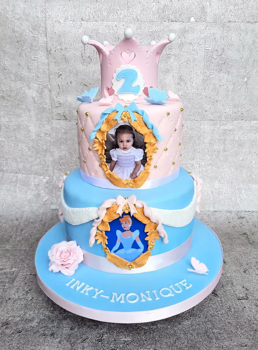 A three-tier birthday cake in pink and light blue. The top tier features a pink crown with the number 2. The middle tier has a quilted design and a framed picture of a young girl. The bottom tier includes a framed image of Cinderella. The light blue base is adorned with a pink ribbon, a pink rose, a butterfly, and the name 'INKY-MONIQUE' in white letters.