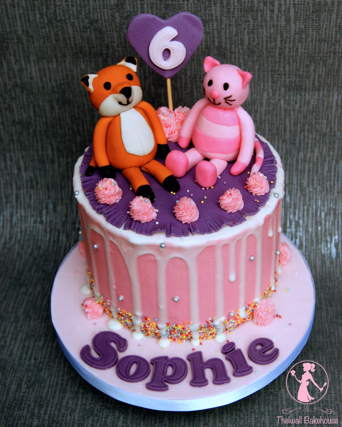 A two-tier birthday cake decorated with fondant figurines of an orange fox and a pink striped cat. The top tier is purple with pink rosettes, and features a purple heart topper with the number 6. The bottom tier is pink with white drips, colorful sprinkles, and silver edible pearls. The light pink cake board has purple letters spelling 'Sophie'.