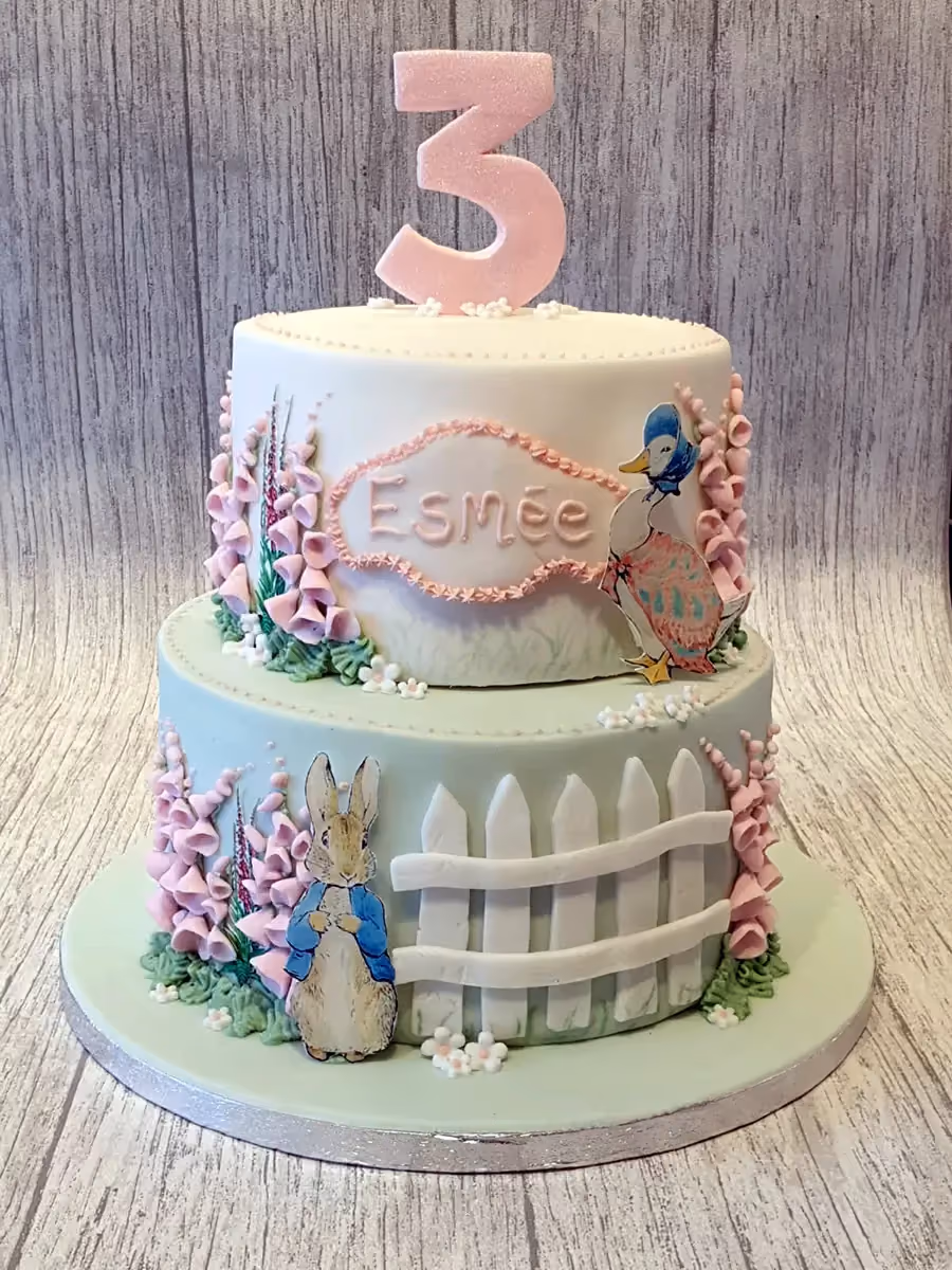 A two-tier Peter Rabbit themed birthday cake with white and pale green fondant, featuring a pink glittery number three on top, the name Esmée, illustrations of Peter Rabbit and Jemima Puddle-Duck, pink foxglove flowers, and a white picket fence.