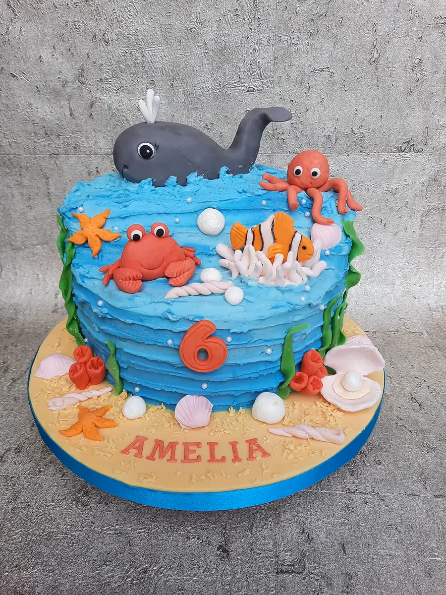An under-the-sea themed birthday cake with blue rippled icing, featuring a gray whale, red crab, orange octopus, and clownfish, adorned with edible coral, starfish, seashells, and pearls, with a red number '6' and the name 'AMELIA' on the sandy base.