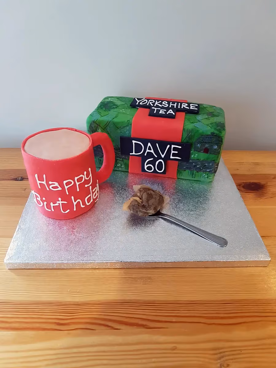 A birthday cake designed as a Yorkshire Tea box and a red mug, displayed on a silver cake board. The tea box cake is green with a red band, featuring 'YORKSHIRE TEA' and 'DAVE 60' in black and white lettering. The red mug cake has 'Happy Birthday' written in white. A spoon with a tea bag sits in front of the cakes.