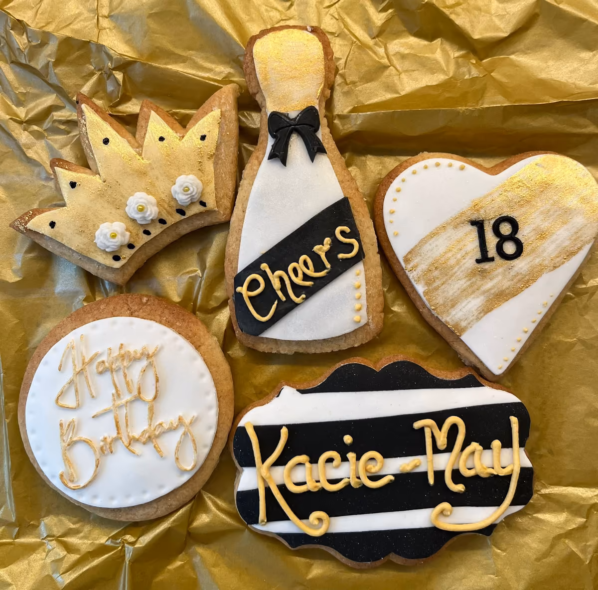 A set of five personalized birthday celebration cookies in gold, black, and white, featuring a crown, a champagne bottle with 'Cheers', a heart with '18', a round cookie with 'Happy Birthday', and a striped plaque with 'Kacie-May'.