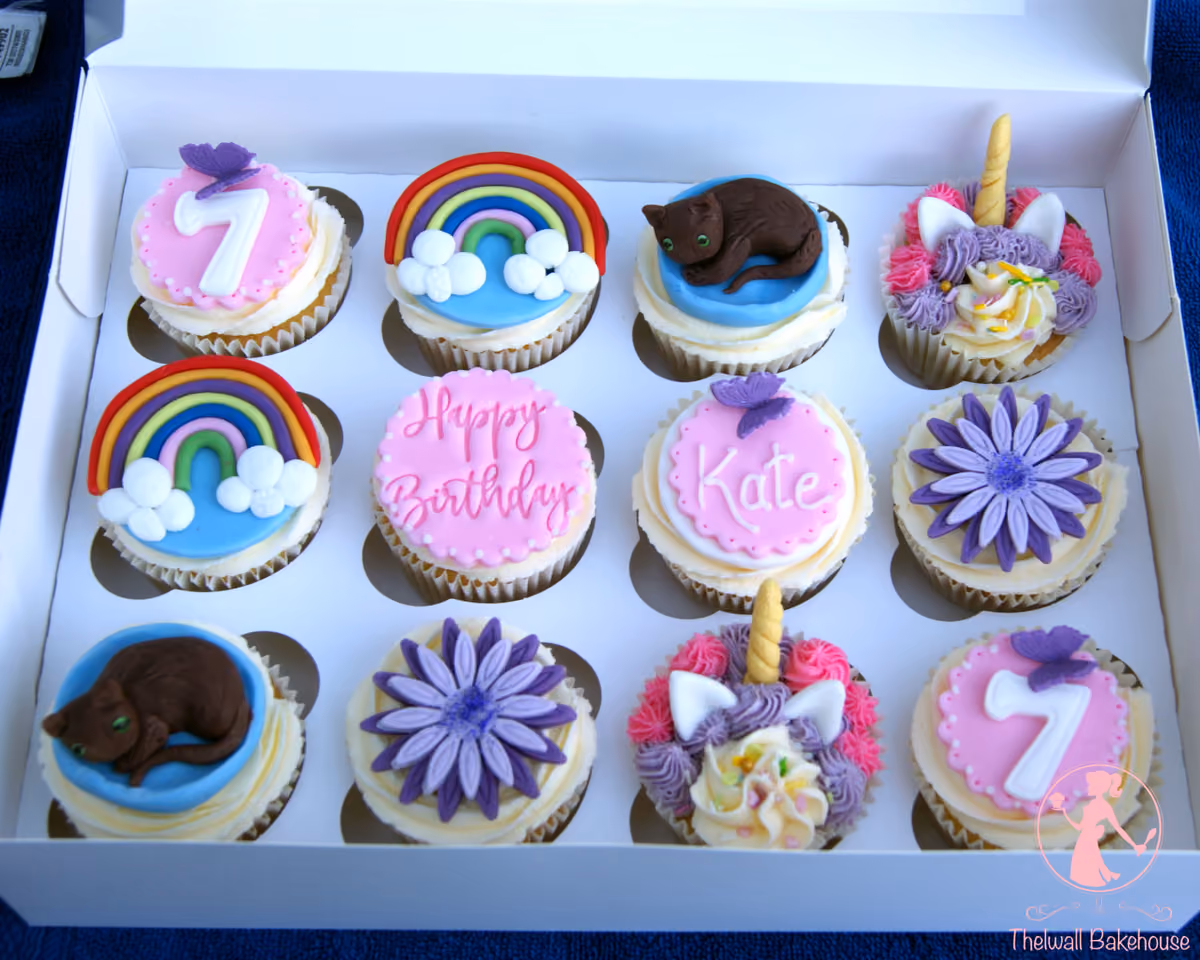A dozen assorted birthday cupcakes in a white box, featuring designs including rainbows, sleeping cats, unicorns, purple flowers, and pink toppers with 'Happy Birthday', 'Kate', and the number '7' with purple butterflies.
