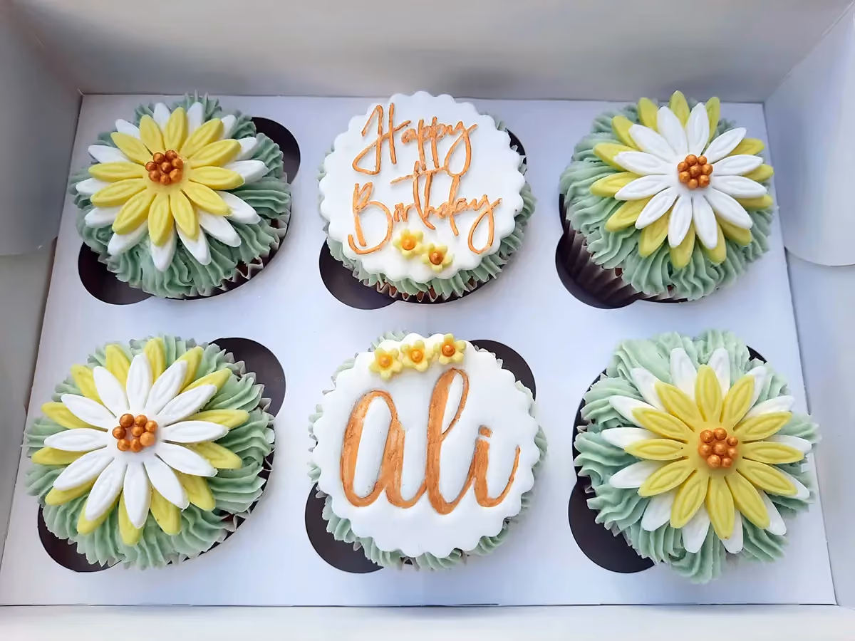 A white box containing six cupcakes. Four cupcakes are decorated with green frosting and layered white and yellow sugar flowers with a gold center. Two cupcakes have white fondant toppers, one inscribed with 'Happy Birthday' in orange script and the other with 'Ali' in orange script, topped with small yellow flowers.