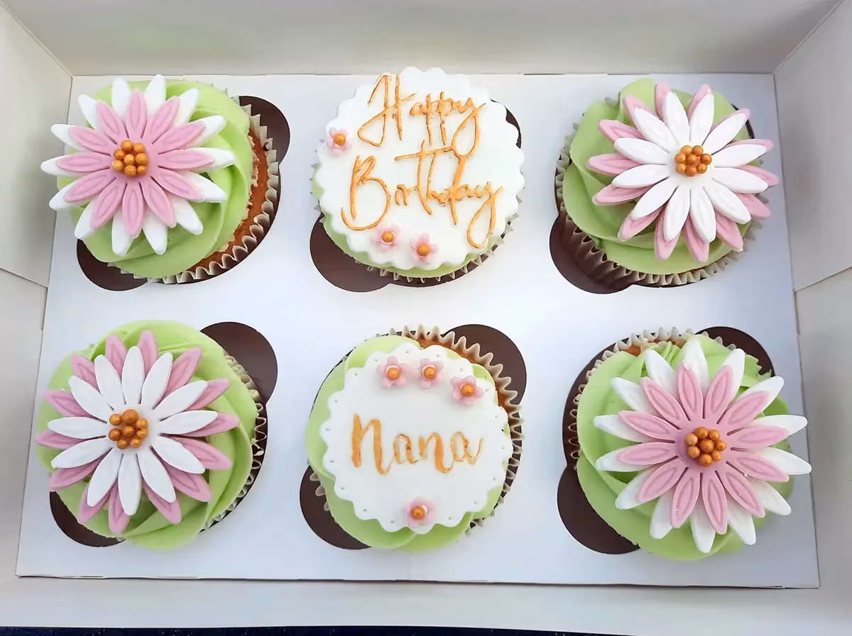 Six cupcakes in a white box with light green frosting. Four cupcakes are topped with layered pink and white sugar flowers, while two have white scalloped fondant toppers, one reading 'Happy Birthday' and the other 'Nana' in gold script, both decorated with small pink flowers.