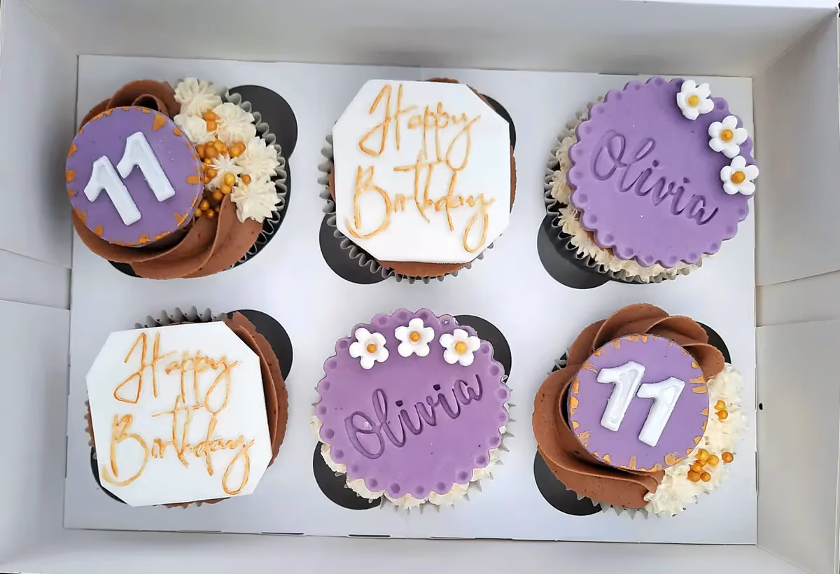Six birthday cupcakes in a white box. Two cupcakes have purple fondant toppers with the name 'Olivia' and small white flowers. Two cupcakes have purple fondant toppers with the number '11' in white, surrounded by gold accents. The remaining two cupcakes have white octagon-shaped fondant toppers with 'Happy Birthday' written in gold script. All cupcakes feature a mix of chocolate and white frosting.