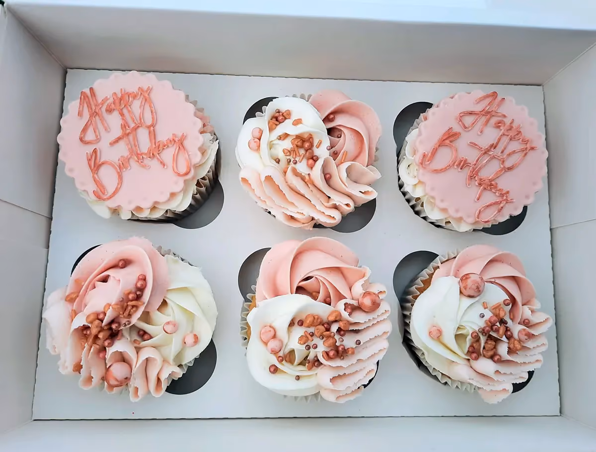 Six birthday cupcakes in a white box, featuring two with pink 'Happy Birthday' toppers and four with pink and white swirled frosting decorated with rose gold sprinkles and edible pearls.