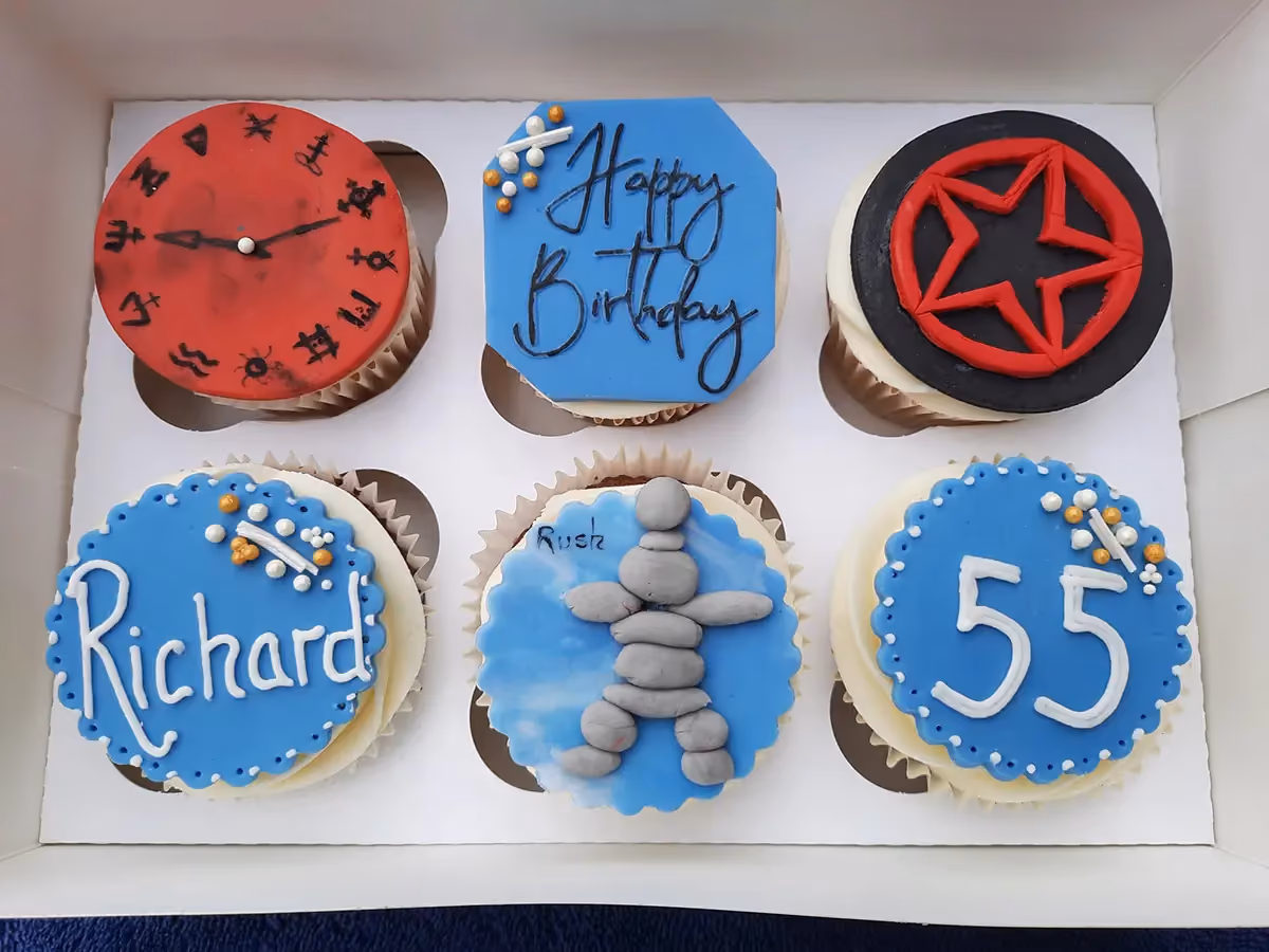 A white box containing six themed birthday cupcakes. The top row features a red clock face with black symbols, a blue octagonal cupcake with 'Happy Birthday' in black script, and a black cupcake with a red five-pointed star. The bottom row shows a blue scalloped cupcake with 'Richard' in white script, a light blue cupcake with a grey Inukshuk figure and 'Rush' written on it, and a blue scalloped cupcake with '55' in white numbers.