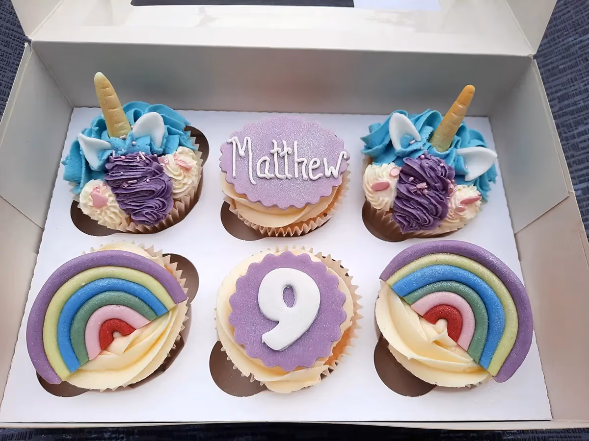 Six birthday cupcakes in a white box, including two unicorn-themed cupcakes, two rainbow-decorated cupcakes, one cupcake with 'Matthew' written on a purple disc, and one cupcake with the number '9' on a purple disc.
