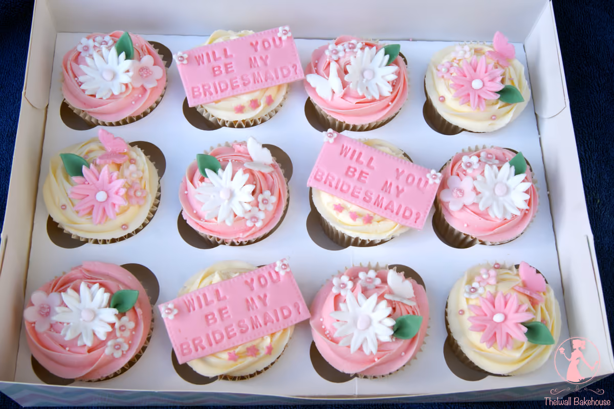 A box of twelve bridesmaid proposal cupcakes, featuring pink and cream frosting, decorated with sugar flowers, butterflies, and three rectangular toppers that read 'WILL YOU BE MY BRIDESMAID?'
