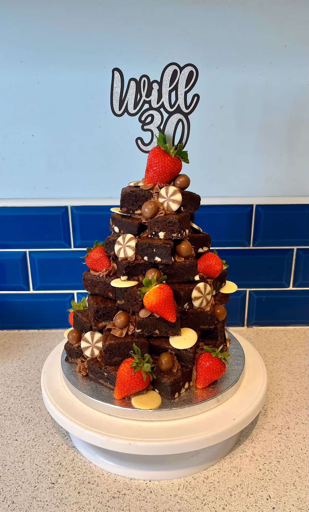 A stacked brownie pyramid cake decorated with fresh red strawberries, chocolate balls, and white and swirled chocolate discs, topped with a 'Will 30' cake topper.