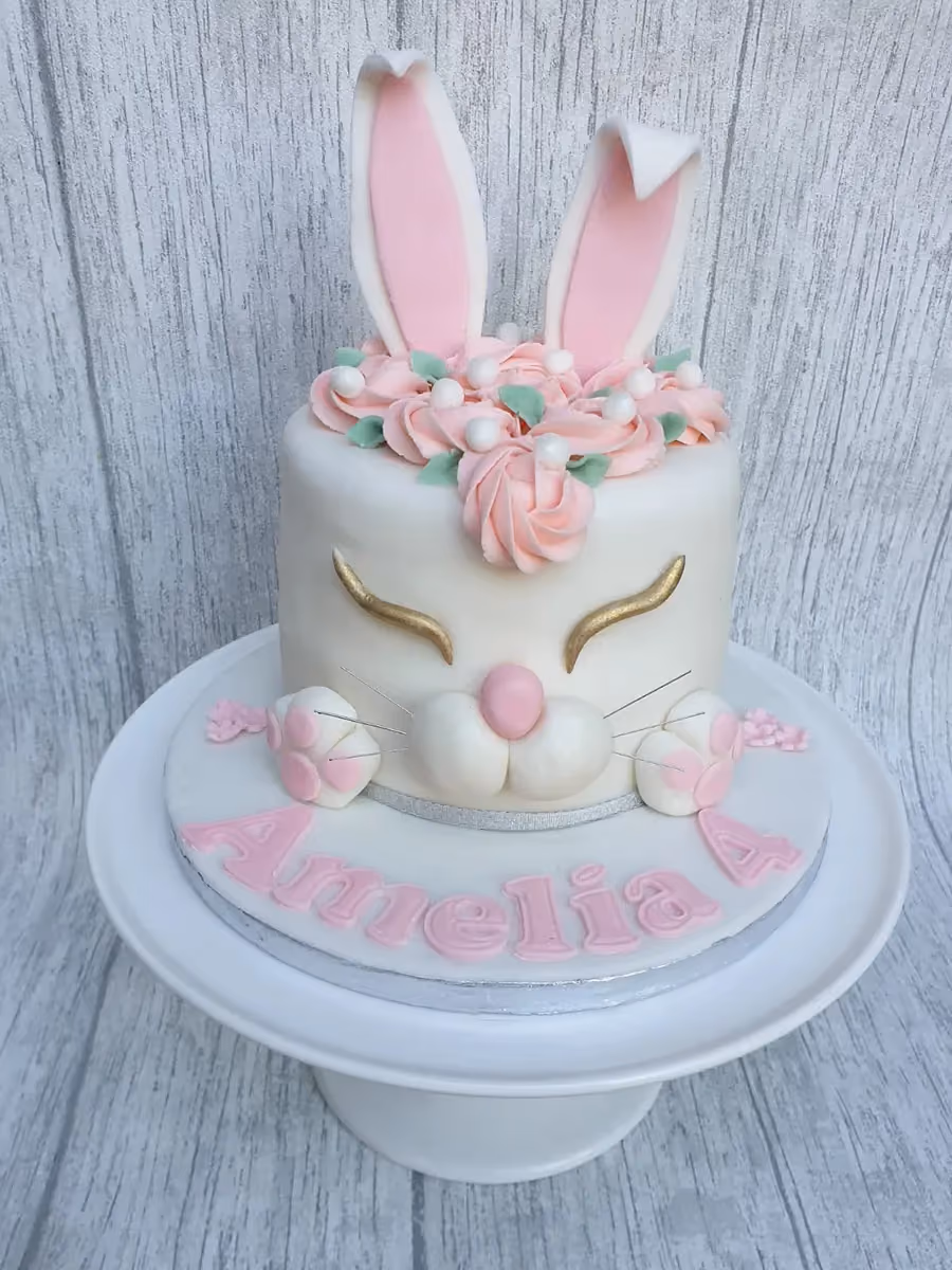 A white bunny-themed birthday cake with tall pink and white ears, pink buttercream rosettes, gold eyelashes, a pink nose, and the name 'Amelia' and number '4' in pink letters on the base.