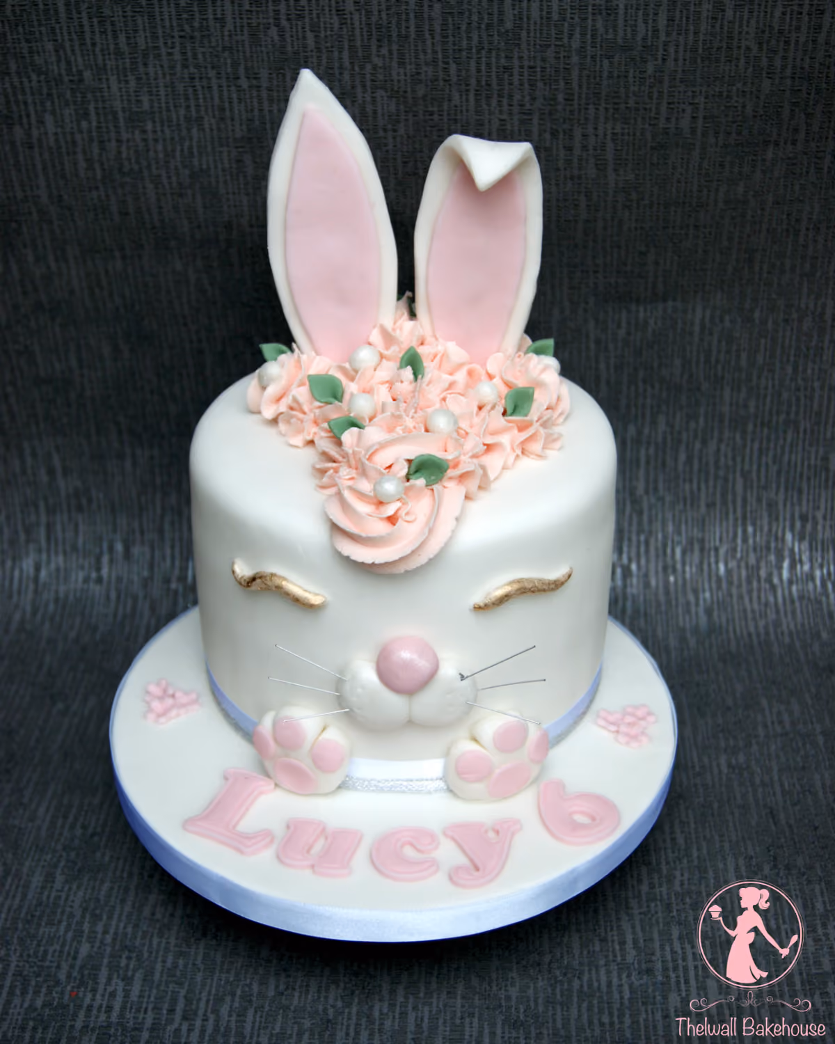 A single-tier white fondant bunny cake with pink inner ears, pink piped frosting flowers and white pearls on top, golden closed eyes, a pink nose, whiskers, and pink bunny paws, with 'Lucy 6' written in pink letters on the cake board.