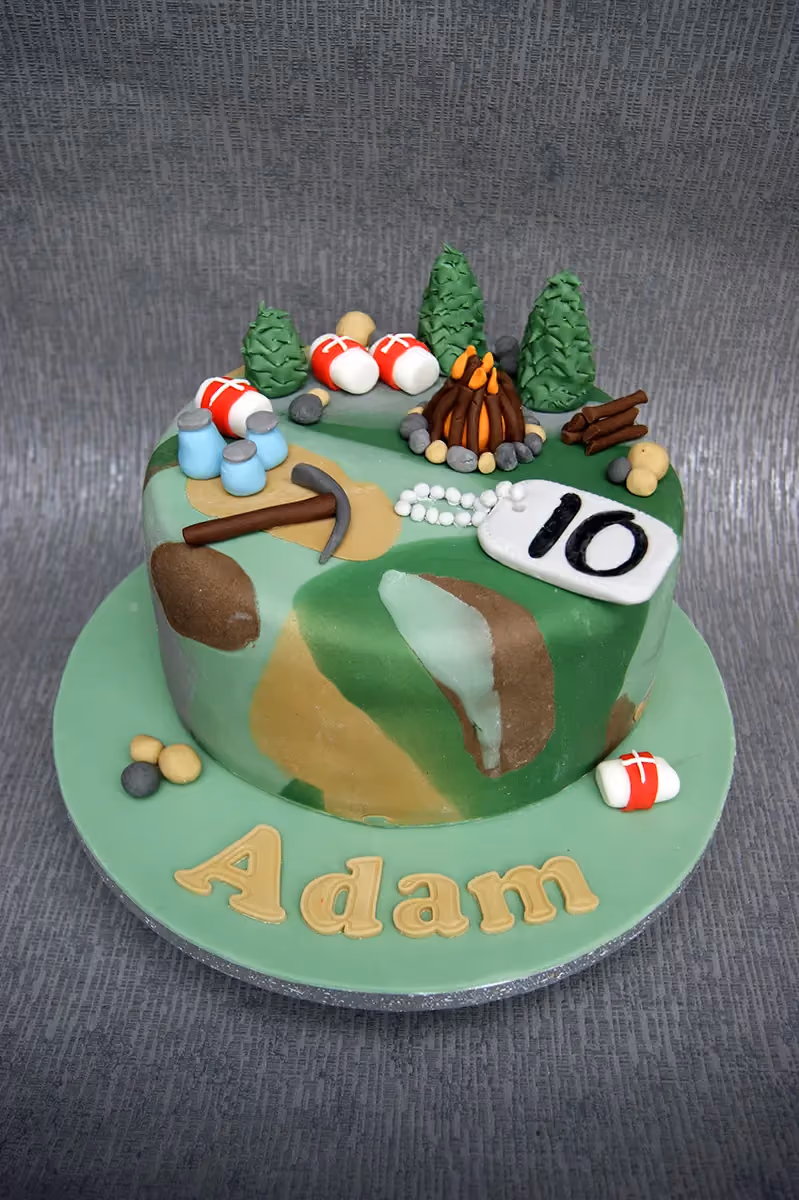 A round birthday cake with a green, brown, and light green camouflage pattern, decorated with edible camping-themed elements including a campfire, green pine trees, blue canteens, a pickaxe, and a white dog tag with the number '10'. The cake sits on a light green board with the name 'Adam' spelled in gold letters.