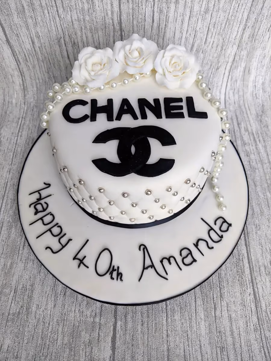 A white Chanel-themed birthday cake featuring black 'CHANEL' text and logo, white sugar roses, and draped pearls. The sides of the cake have a quilted pattern with silver edible beads. The cake board reads 'Happy 40th Amanda' in black script.