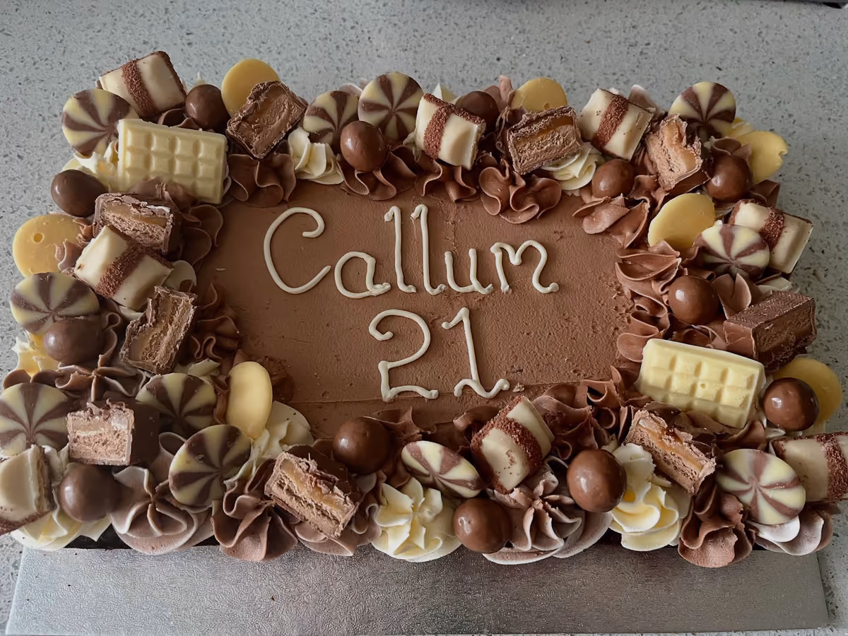 Rectangular chocolate birthday cake for 'Callum 21', decorated with an assortment of chocolates including Kinder Bueno, white chocolate bars, chocolate malt balls, and swirled chocolate discs, surrounded by chocolate and white frosting swirls.