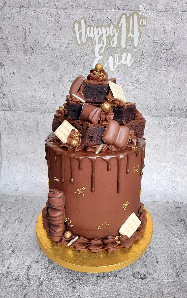 A tall, single-tier chocolate drip cake covered in a cascade of chocolate brownies, various milk and white chocolates, and gold sprinkles, with a 'Happy 14th Eva' cake topper at the top.