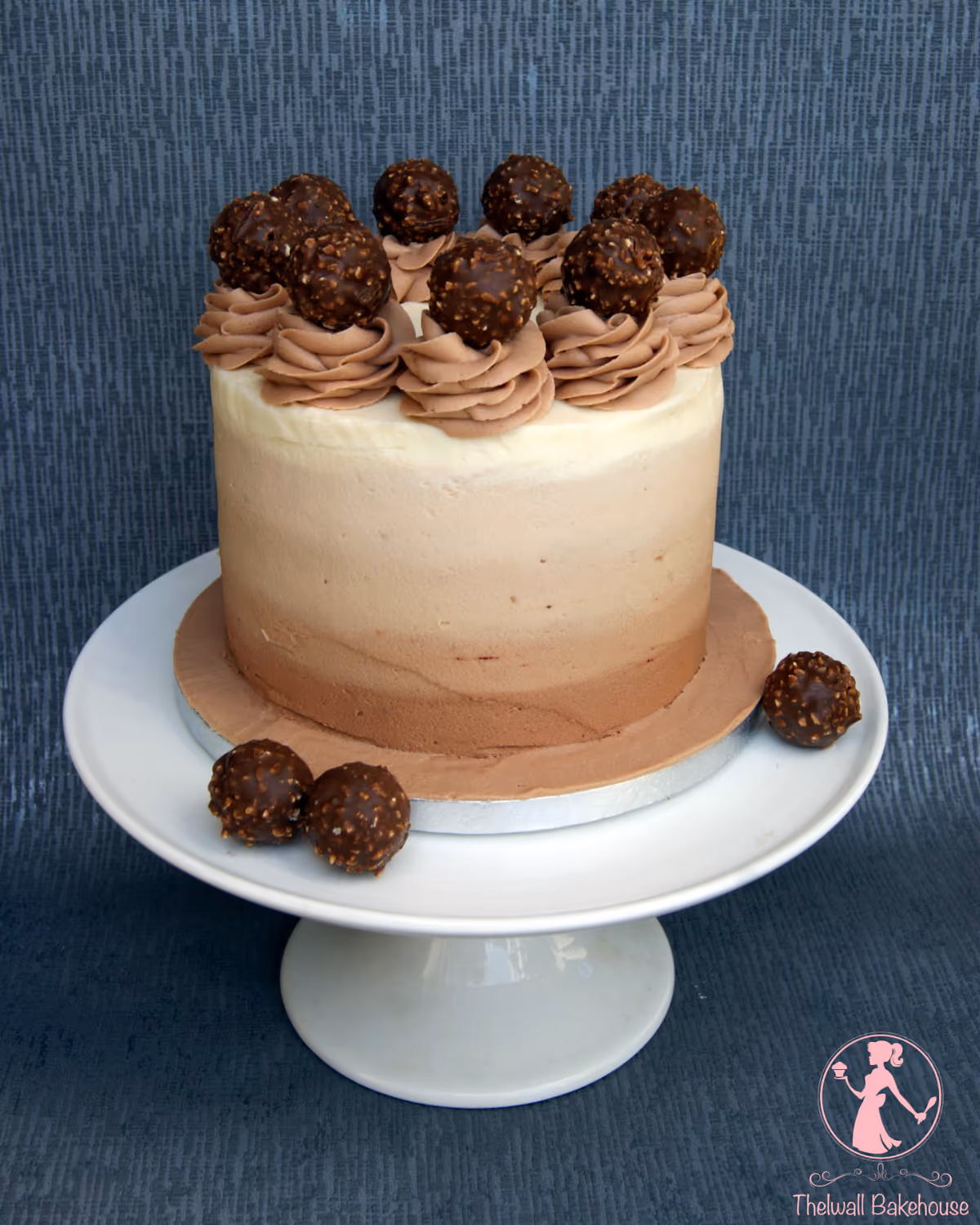 A round cake with ombre frosting, transitioning from white at the top to light brown and then to dark brown at the base, decorated with swirls of brown buttercream and several chocolate-hazelnut candies on top, presented on a white cake stand with a few extra candies.