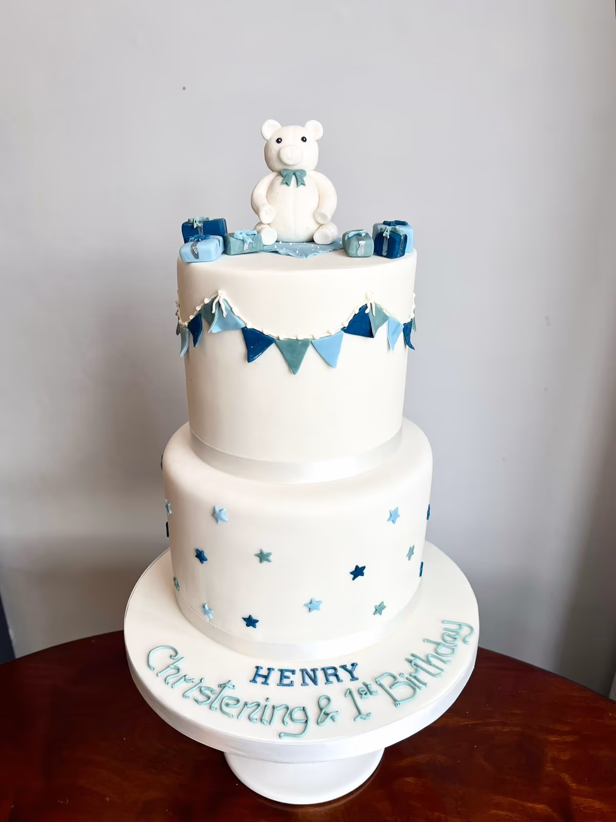 A two-tier white fondant cake for a boy's Christening and 1st Birthday, decorated with a white teddy bear, blue and green gift boxes and bunting on the top tier, and blue and green stars on the bottom tier. The cake board is inscribed with 'HENRY Christening & 1st Birthday'.