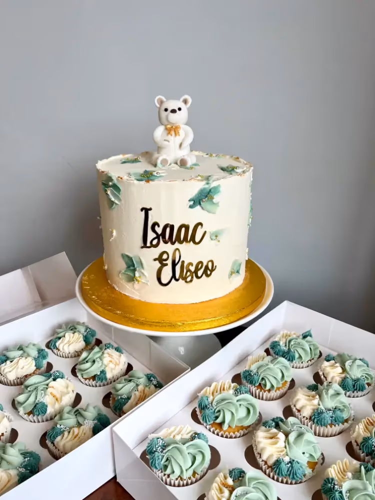 A white buttercream cake decorated with green leaf accents and a white teddy bear topper, featuring the names 'Isaac Eliseo' in gold script, displayed with two boxes of matching green and white frosted cupcakes.