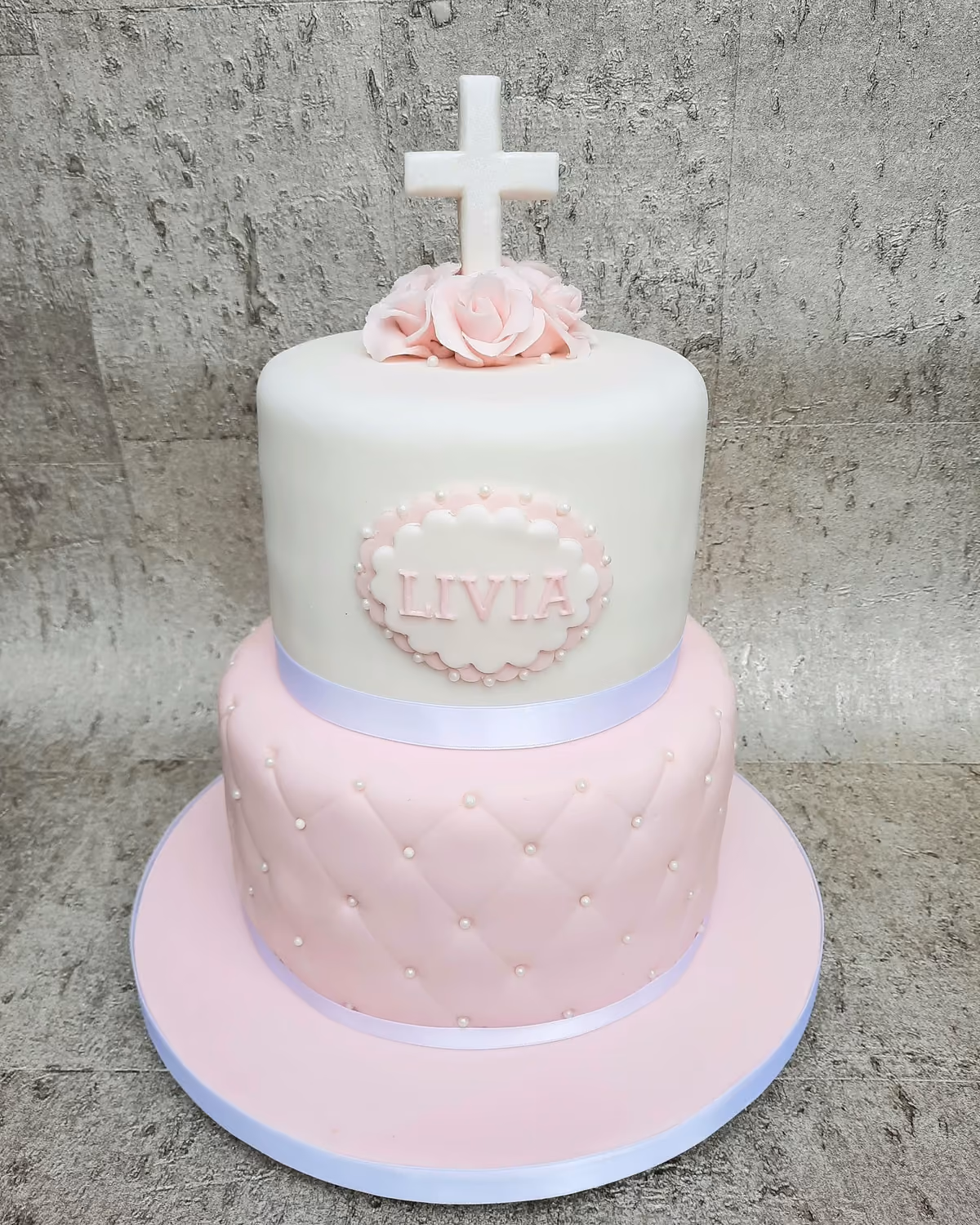 A two-tier christening cake for a girl, featuring a white top tier with a white cross, pink sugar roses, and a pink and white plaque with the name 'LIVIA', above a pink quilted bottom tier adorned with white pearls. The cake sits on a pink cake board.