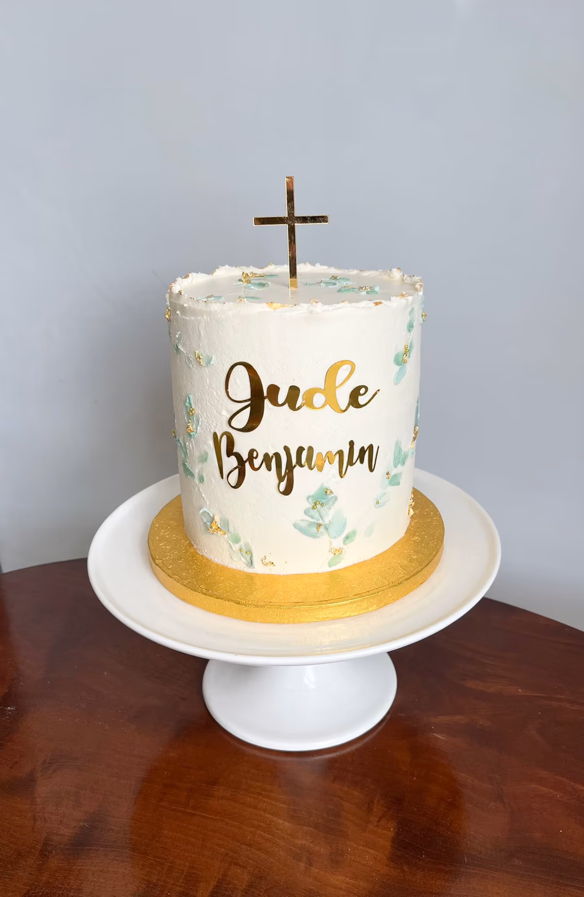 A white single-tier christening cake for Jude Benjamin, decorated with painted green leaves, gold flecks, and topped with a golden cross.